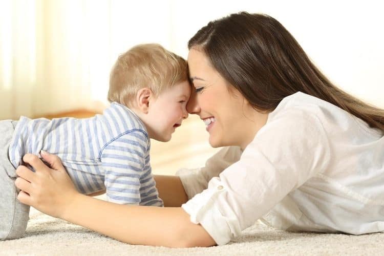 Mom and toddler facing each other stomachs on ground with heads ttouching laughing playfully