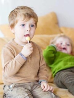 Toddler holding a sucker sitting on a couch with a sibling in the back also on couch