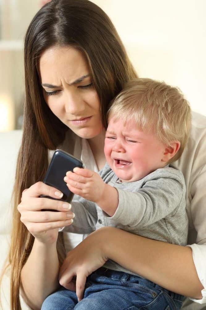 Mom playing with a smartphone while toddler is in her lap crying